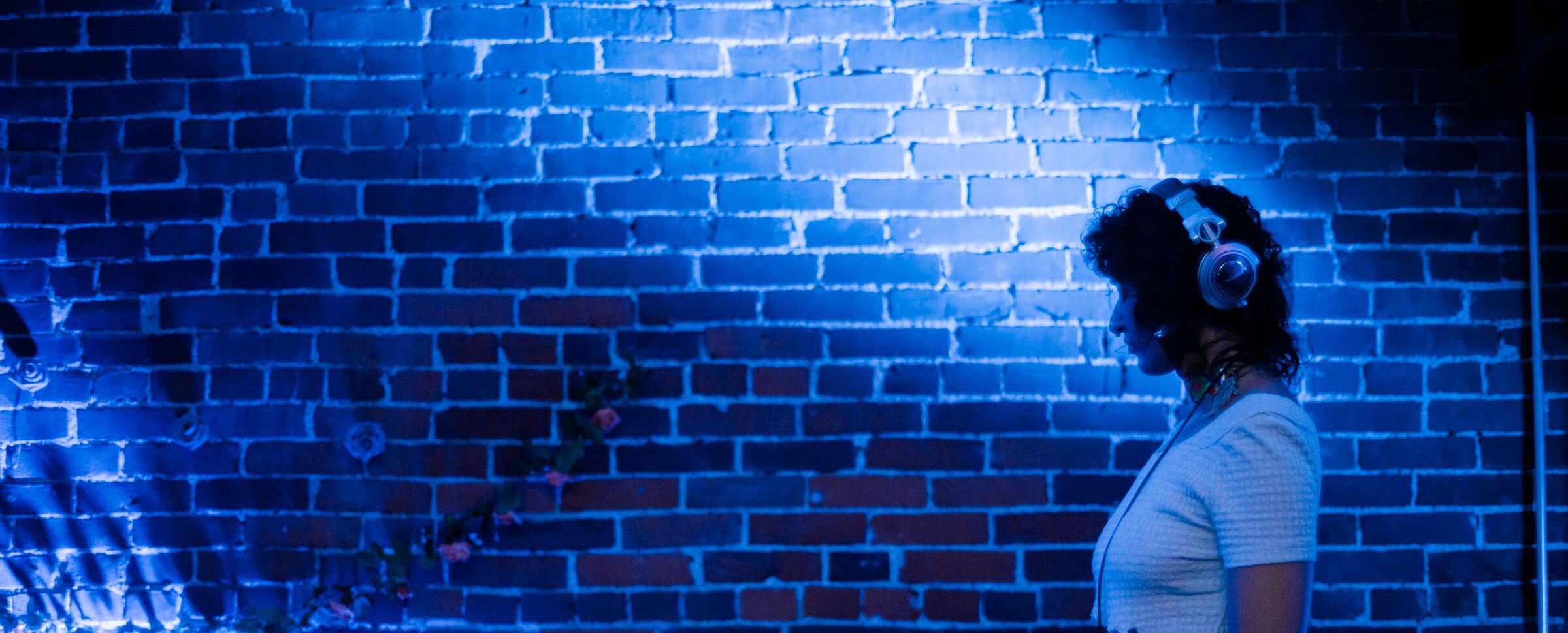 A profile of Regina Martinez wearing headphones, standing in front of a blue-lit brick wall, with a few flowers opposite her